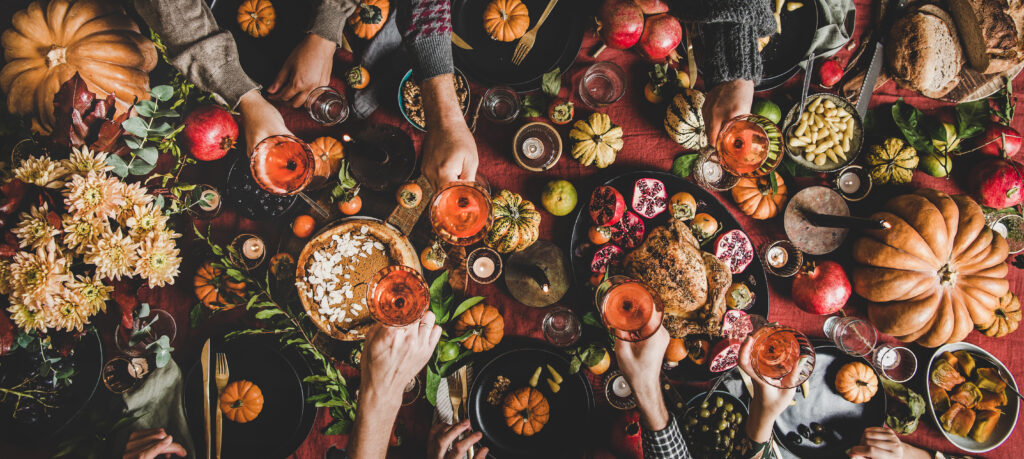 Family celebrating at Thanksgiving table. Flat-lay of feasting peoples hands with glasses of wine over Friendsgiving table with Autumn food, candles, roasted turkey and pumpkin pie over wooden table