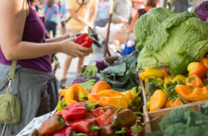 Colorful fresh produce at a farmers market in the foreground with a woman and her purchases in the background.