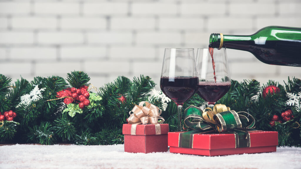 Red wine is pouring from bottle in to glass decorating with red gift boxes and Christamas ornaments. Copy space at top area with blurred white brick wall.